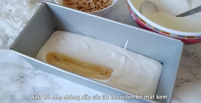 Lót một lớp kem xuống đáy khuôn bánh mì. Xếp lát chuối lên trên, phủ kín bề mặt kem.