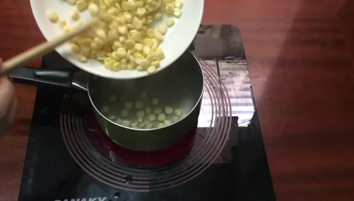 Luộc hạt ngô với 100g đường trong nước sôi khoảng 1 phút, sau đó tắt bếp và vớt ngô ra ngâm vào nước lạnh cho giòn.